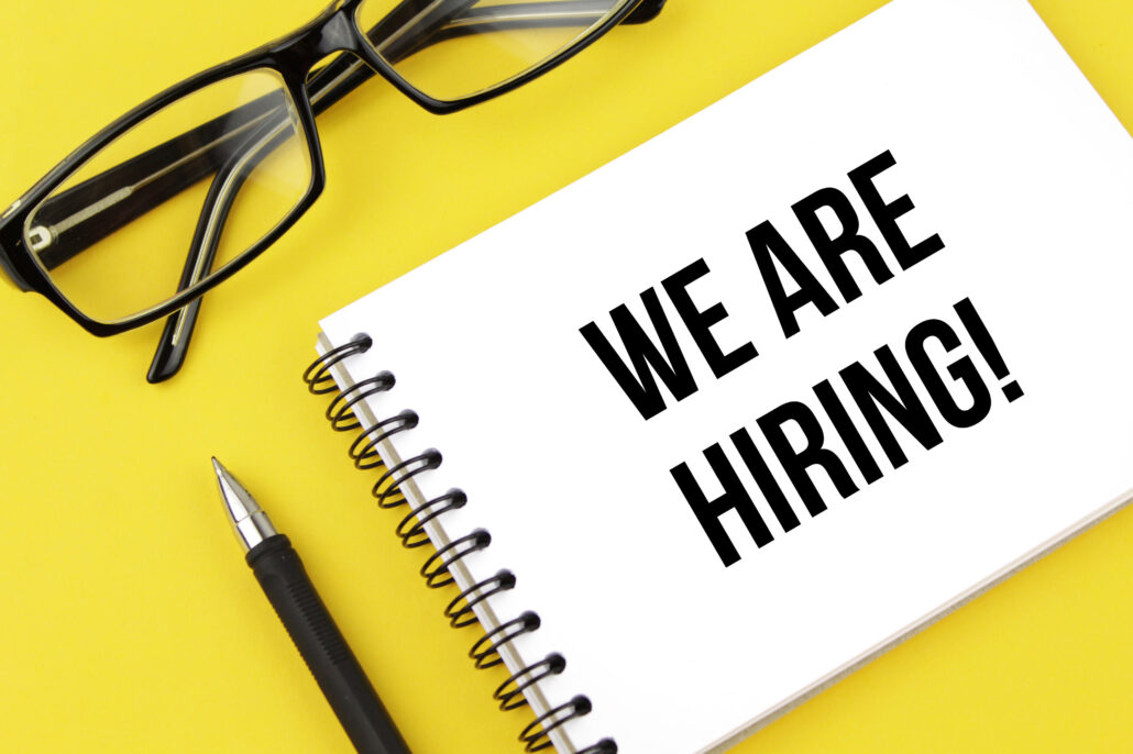 yellow background with black pen, black glasses, and white notepad with the words WE ARE HIRING!