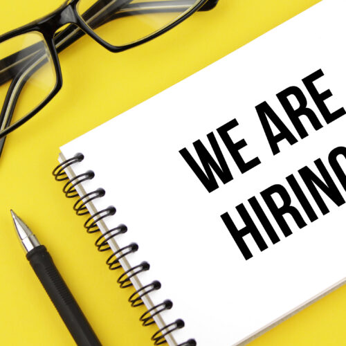yellow background with black pen, black glasses, and white notepad with the words WE ARE HIRING!