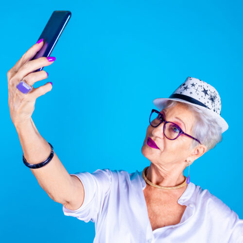 Stylish senior woman influencer (Granfluencer) fashionable taking selfie or streaming live on blue background enjoying social trends - vlogger, technology, vanity concept