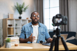 African American man in denim shirt and eyeglasses sitting at table, gesturing and talking while recording video on camera. Male blogger doing live stream in his social network.