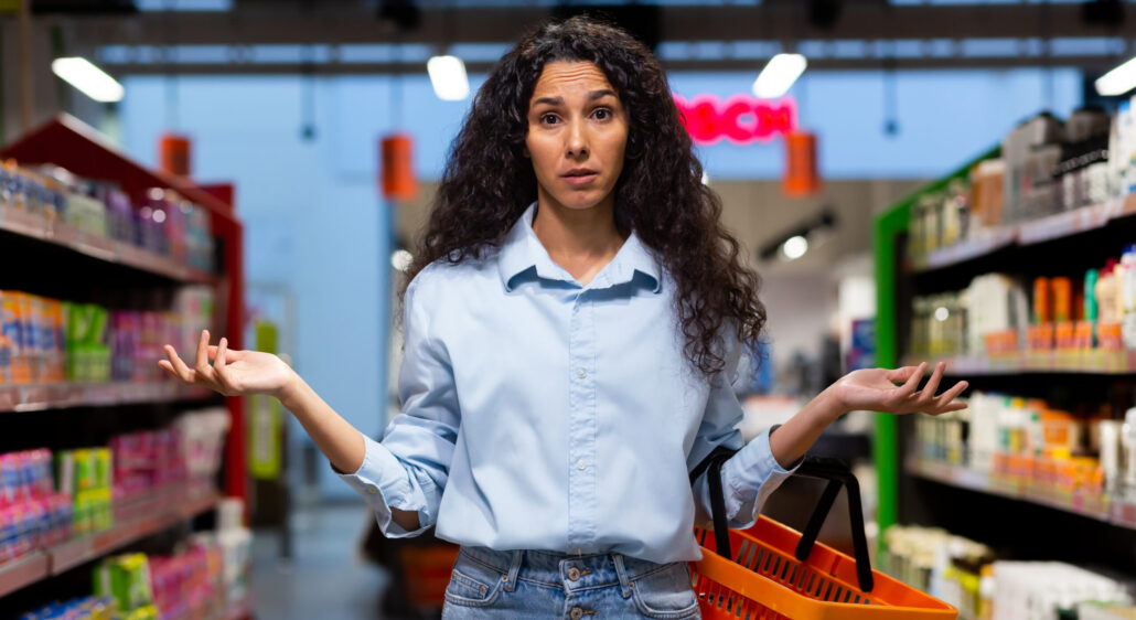 A worried young Latin American woman stands in a supermarket between the shelves with goods, holds a basket, spreads her hands, looks at the camera. Signifying broken branding and confused customers