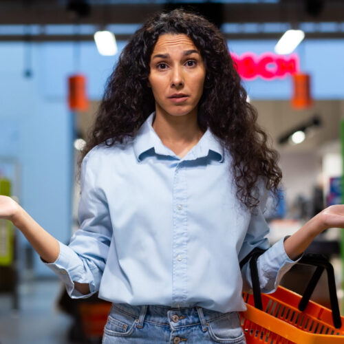 A worried young Latin American woman stands in a supermarket between the shelves with goods, holds a basket, spreads her hands, looks at the camera. Signifying broken branding and confused customers