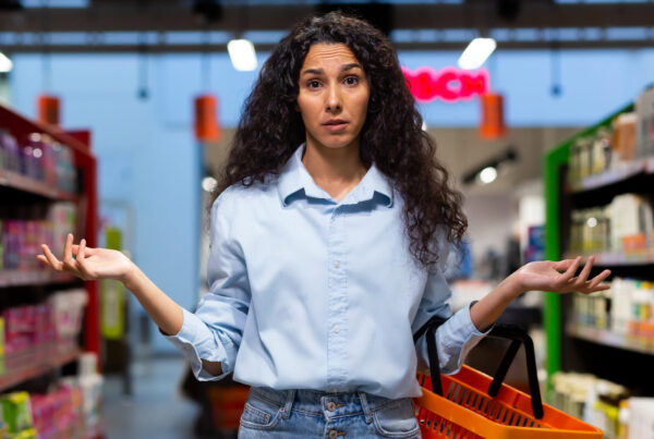 A worried young Latin American woman stands in a supermarket between the shelves with goods, holds a basket, spreads her hands, looks at the camera. Signifying broken branding and confused customers