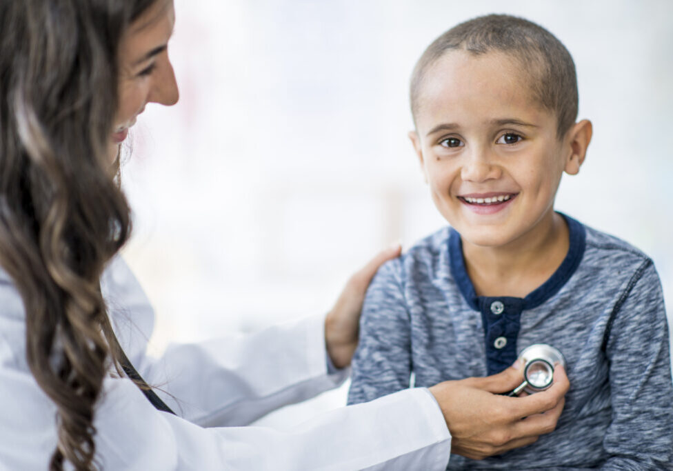 An Ethnic female doctor is indoors in a hospital room. She is wearing medical clothing and a stethoscope. She is checking an Ethnic boy's heart rate.